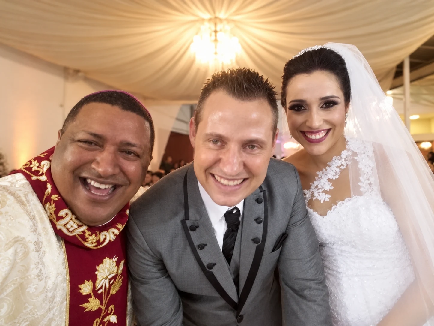 Celebrante e casal de noivos sorrindo em uma fotografia próxima, sob um teto decorado com tecido.
